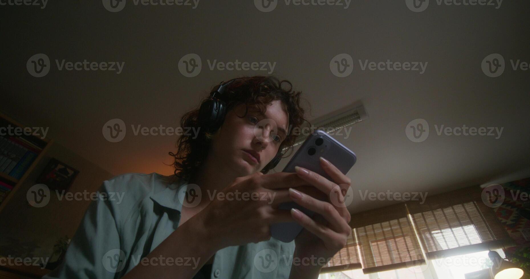A young woman with headphones uses a mobile phone sitting on a bed at home with a sad look photo