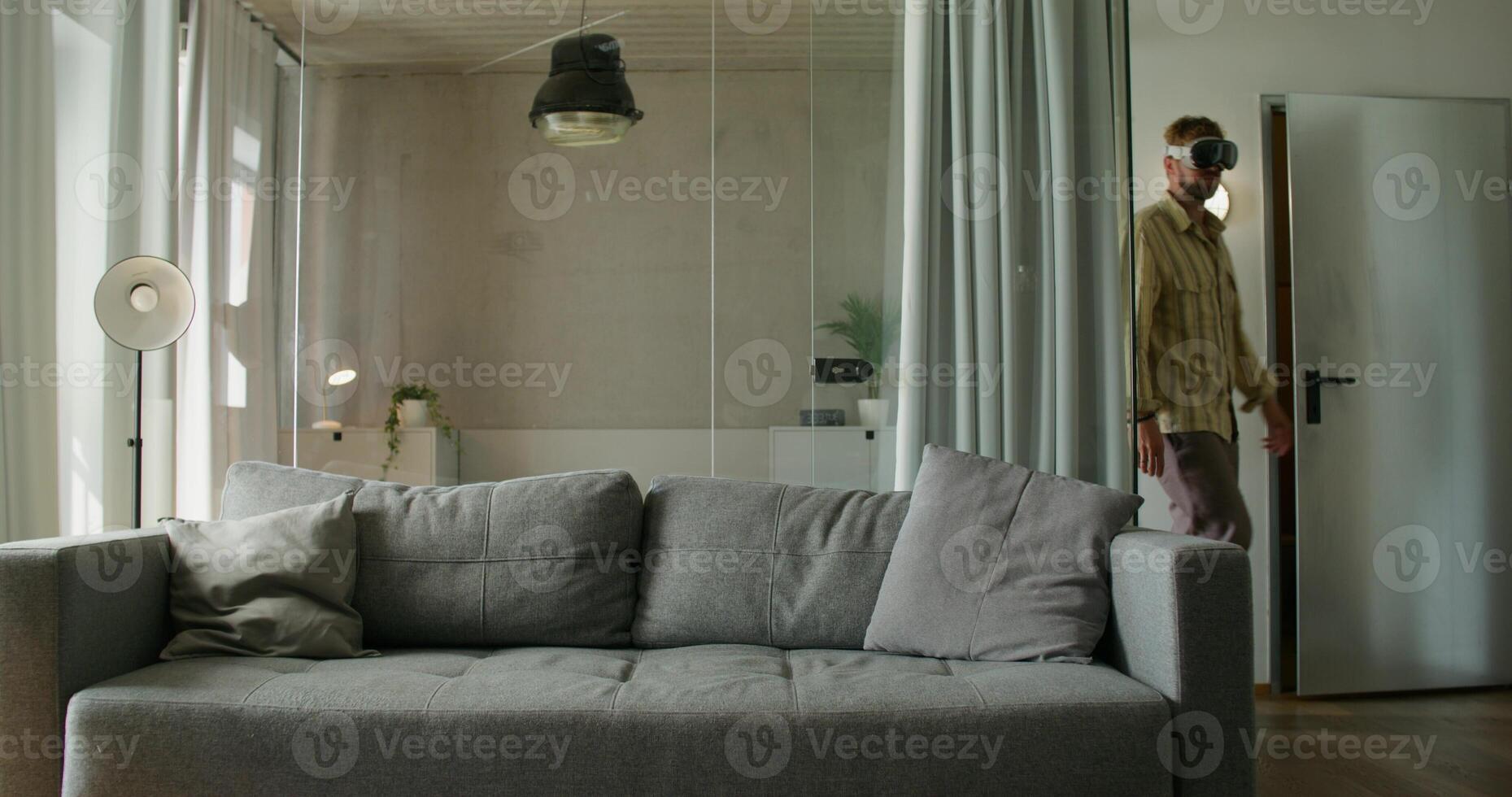 A man in a virtual reality headset sits down on the sofa and starts moving his hands. Daylight, home interior photo