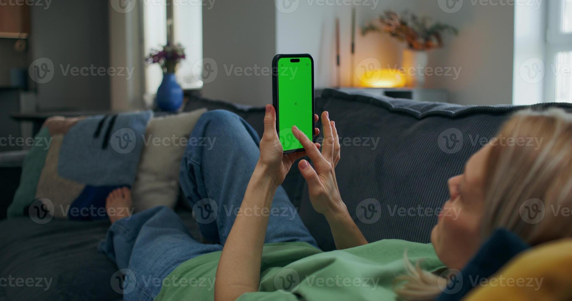 A woman uses a mobile phone with green screen lying on the couch at home. An unrecognizable person photo