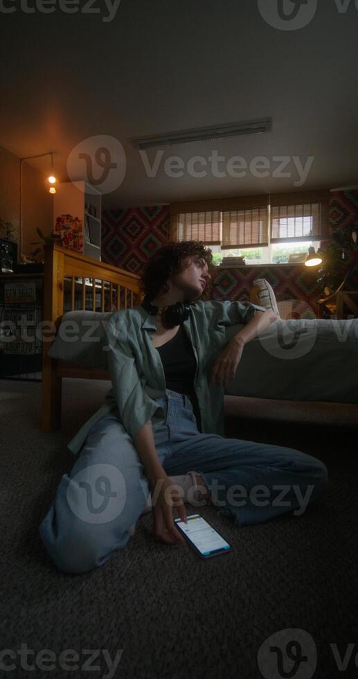 A young woman texting messages on a mobile phone while sitting on the floor near the bed with a depressed look photo