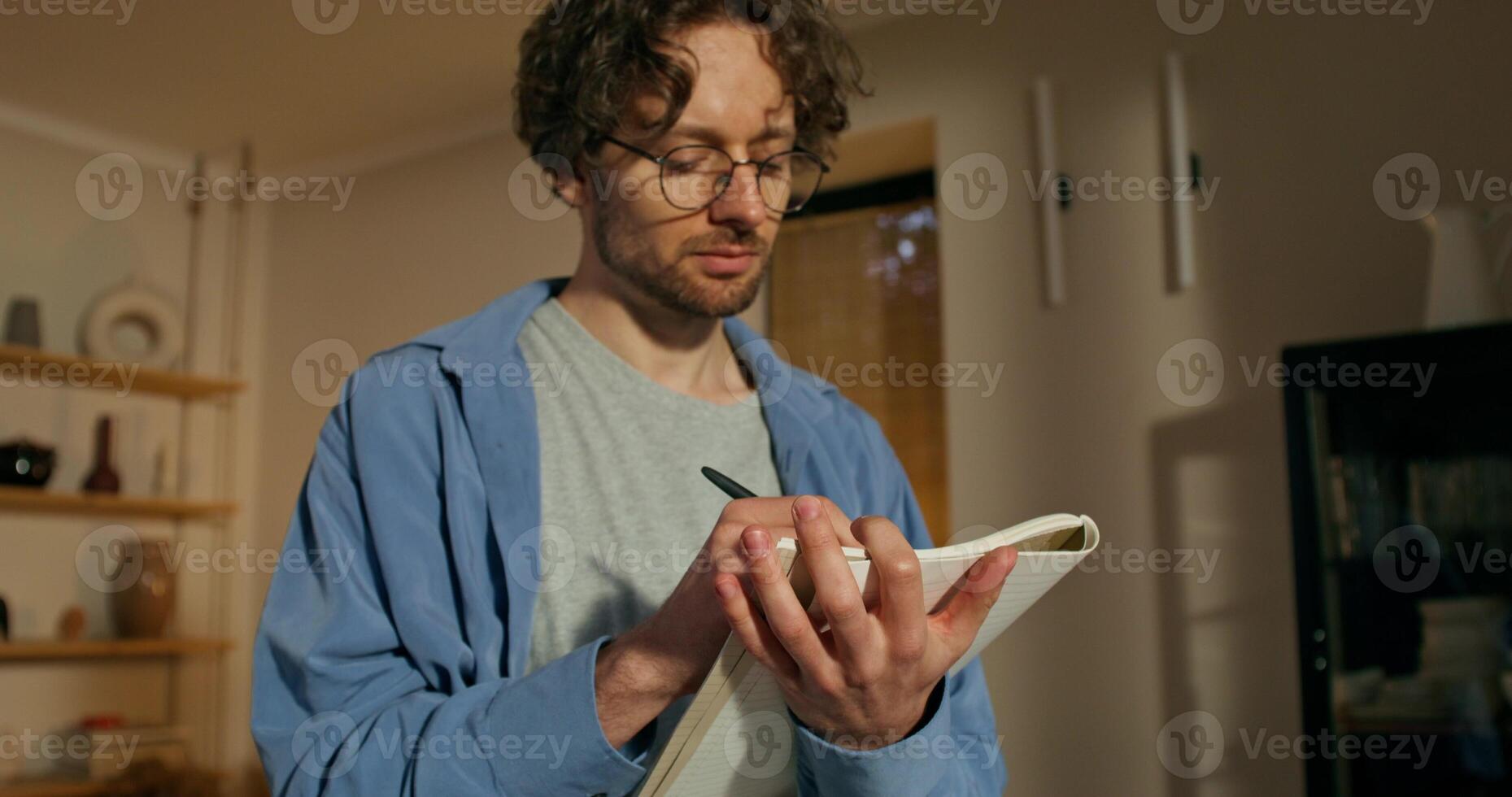 A man with glasses writes in a notebook standing in a stylish home interior with a thoughtful look photo