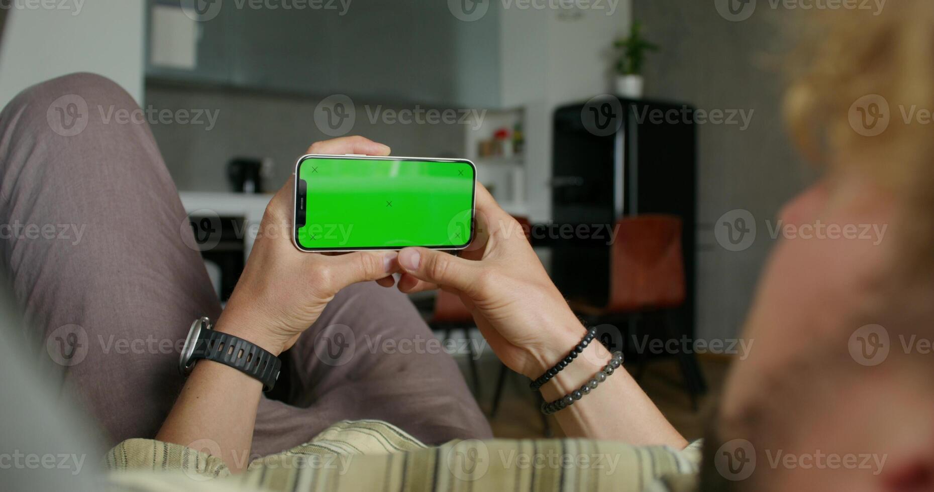 A man holds a mobile phone with a green screen lying on the couch at home. Close-up of his hands, an unrecognizable person photo