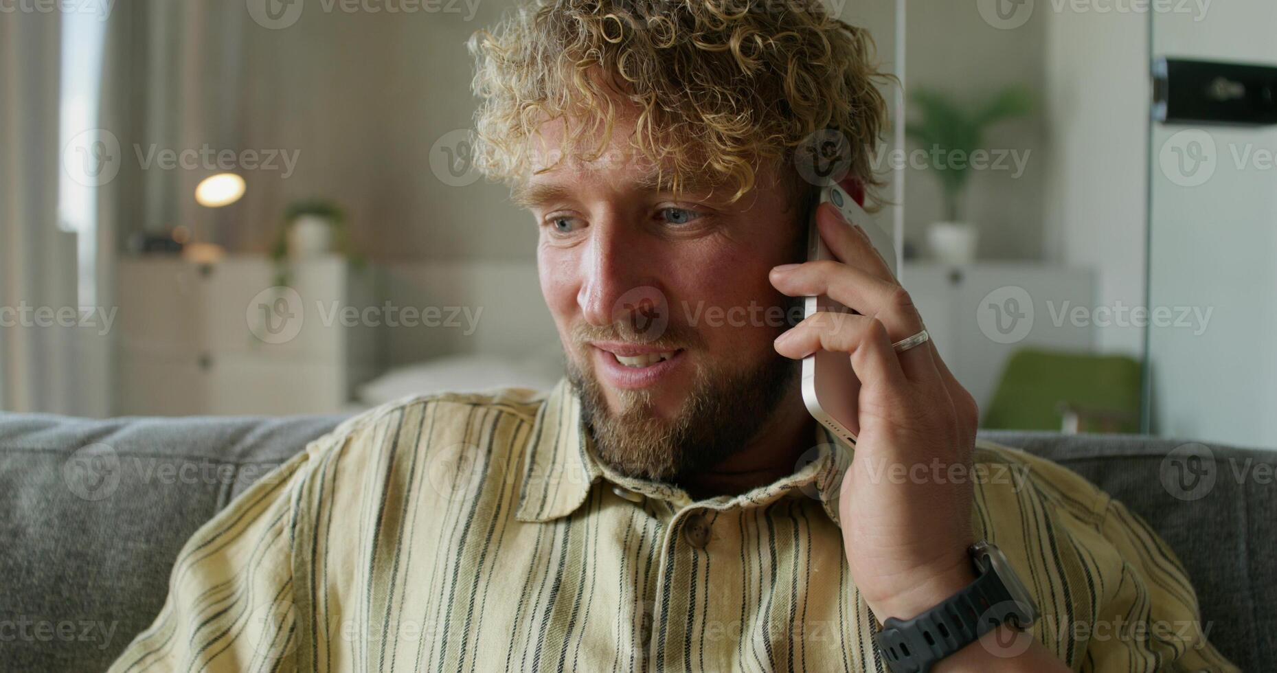 A bearded man is talking on a mobile phone sitting on the couch at home. Close-up of his smiling face. Daylight photo
