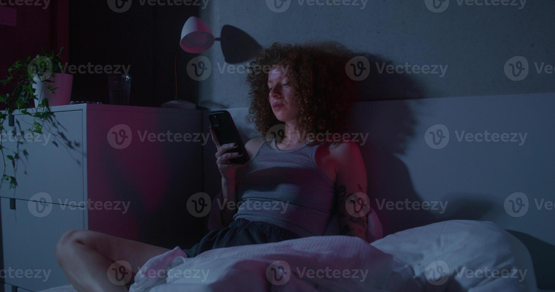 A woman is typing on a mobile phone while sitting on the bed at night. Dark room, home interior photo