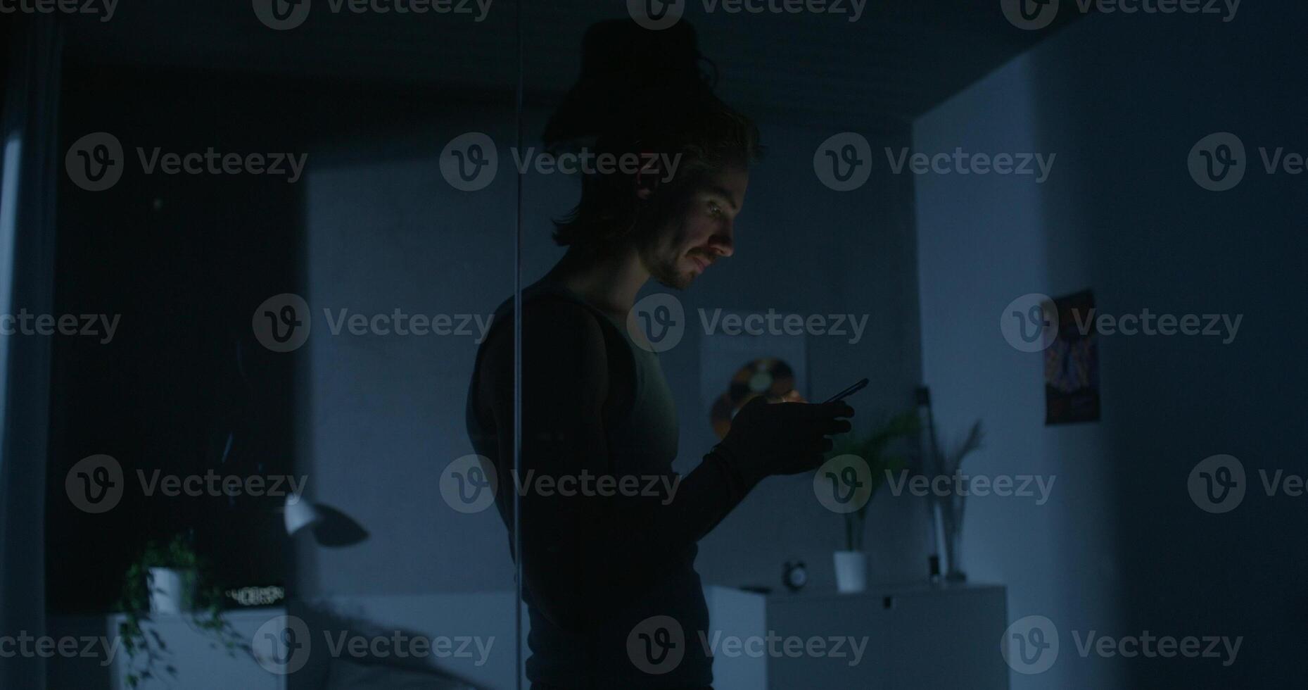 A man uses a mobile phone while walking around the bedroom at night. His wife is sleeping in bed. Dark room, home interior photo