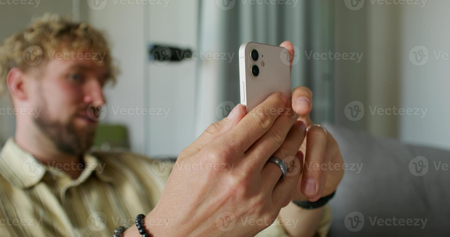 A bearded man in casual clothes is talking on a chat on a mobile phone sitting on the couch at home. The focuses on his hands. Daylight photo