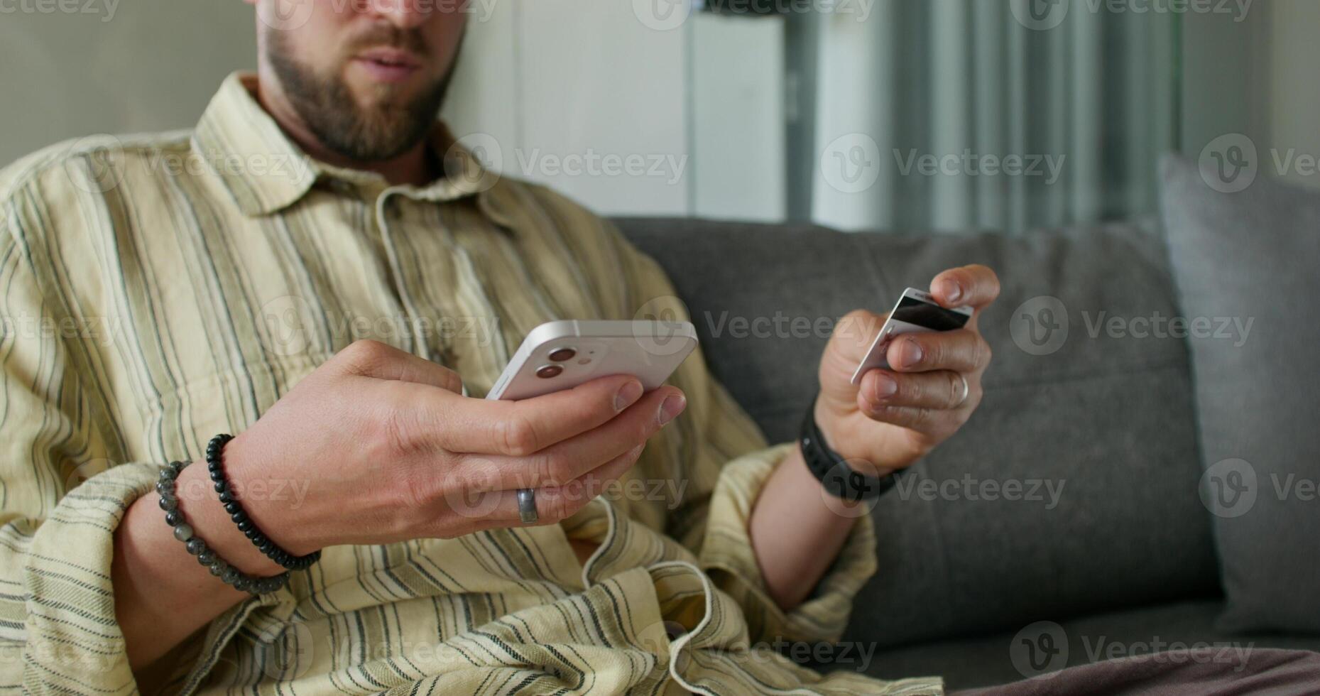 A man enters credit card details into a mobile phone for online purchases while sitting on the couch at home. Close-up of his hands, an unrecognizable person photo