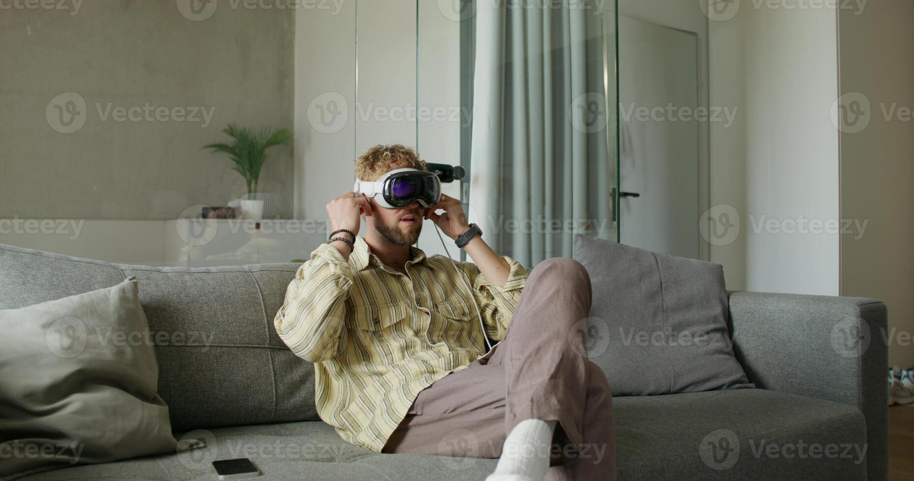 A man in a virtual reality headset puts on earphones and starts a chat sitting on the couch at home. He smiles, talks and moves his arms. Daylight photo
