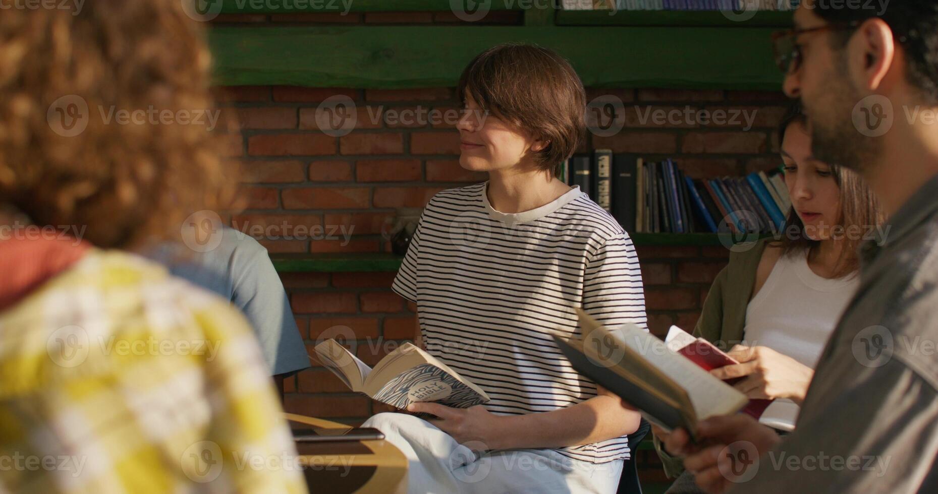 Group of young people are smiling and talking while sitting at a table indoors. They are holding Bibles in their hands. Daylight photo