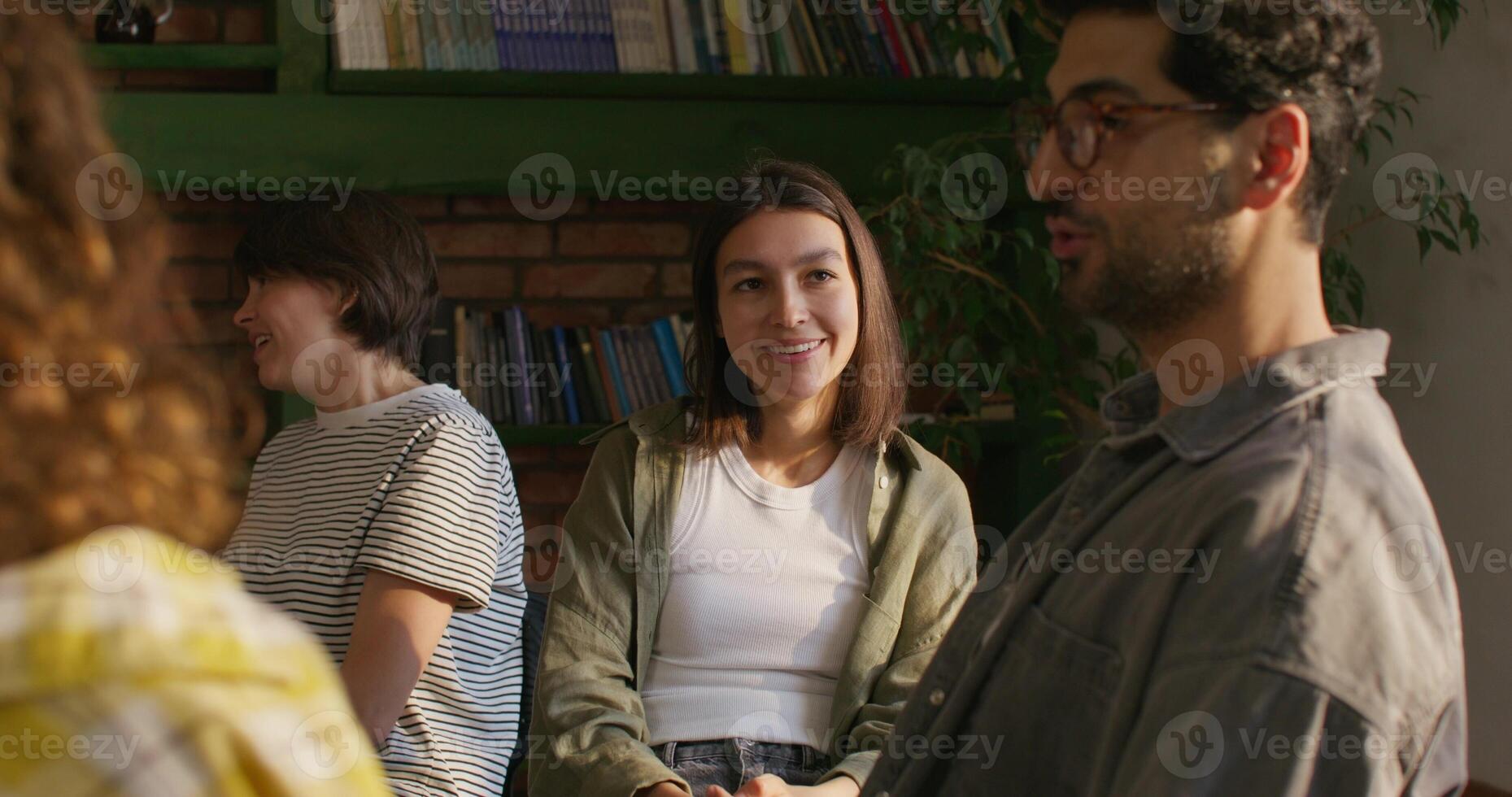 Young people in casual clothes smile and talk while sitting at a table indoors. Daylight photo