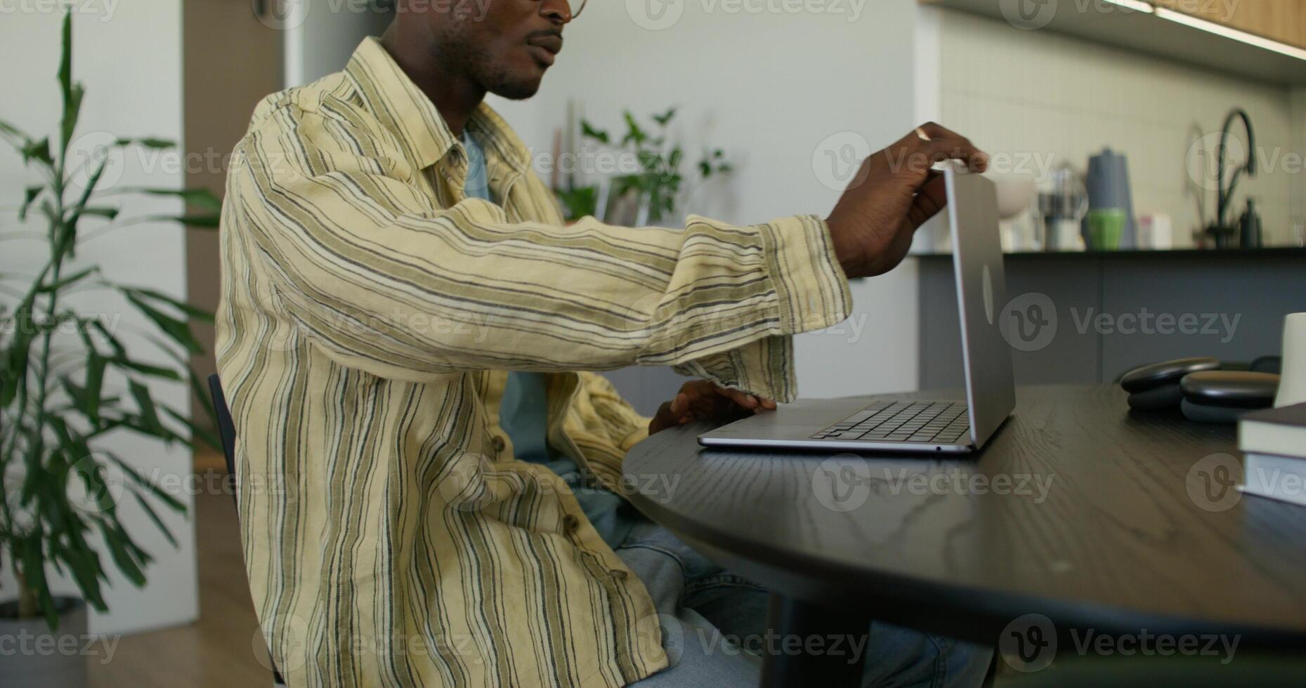 A man in casual clothes opens a laptop and starts typing while sitting at a table at home with a serious look. Daylight photo