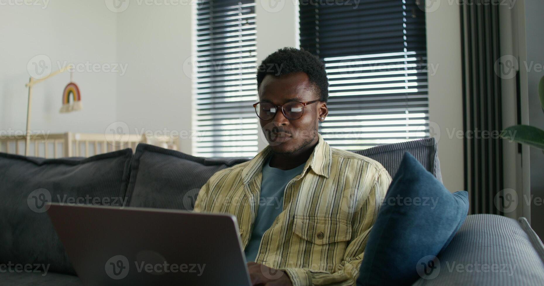 A man in glasses and casual clothes uses a laptop while sitting on the couch at home with a serious look. Daylight photo