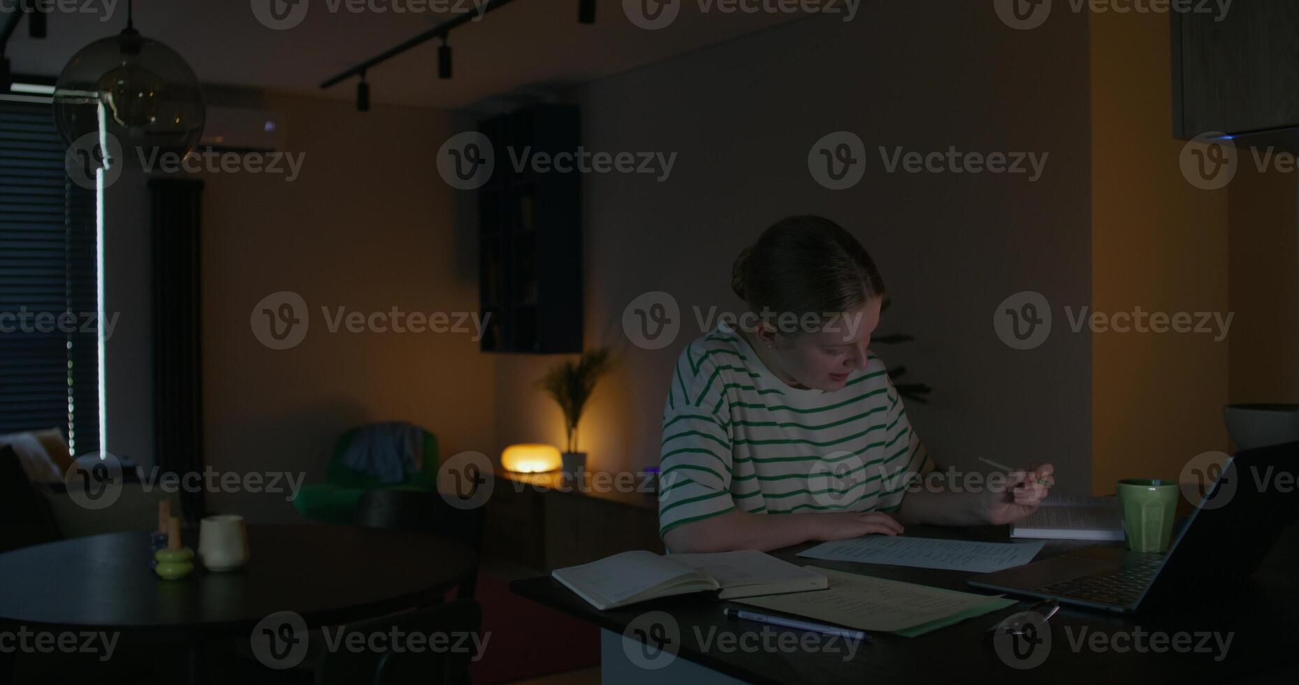 A young woman is talking on chat and writing on paper while sitting at a table in the dark. Online exam photo