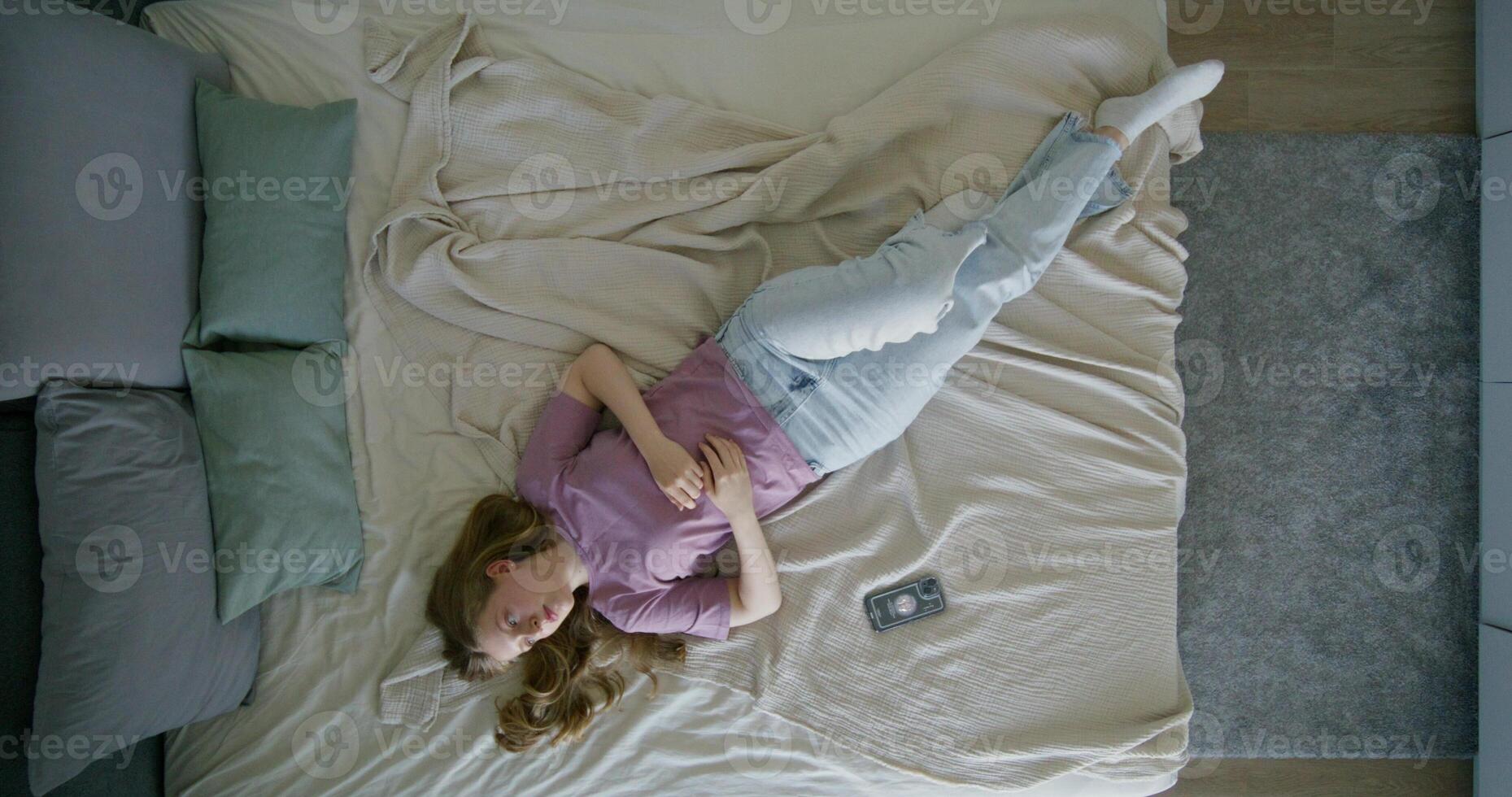 A young woman in casual clothes is lying on a bed during the day. The mobile phone is next to her. Top view, daylight photo
