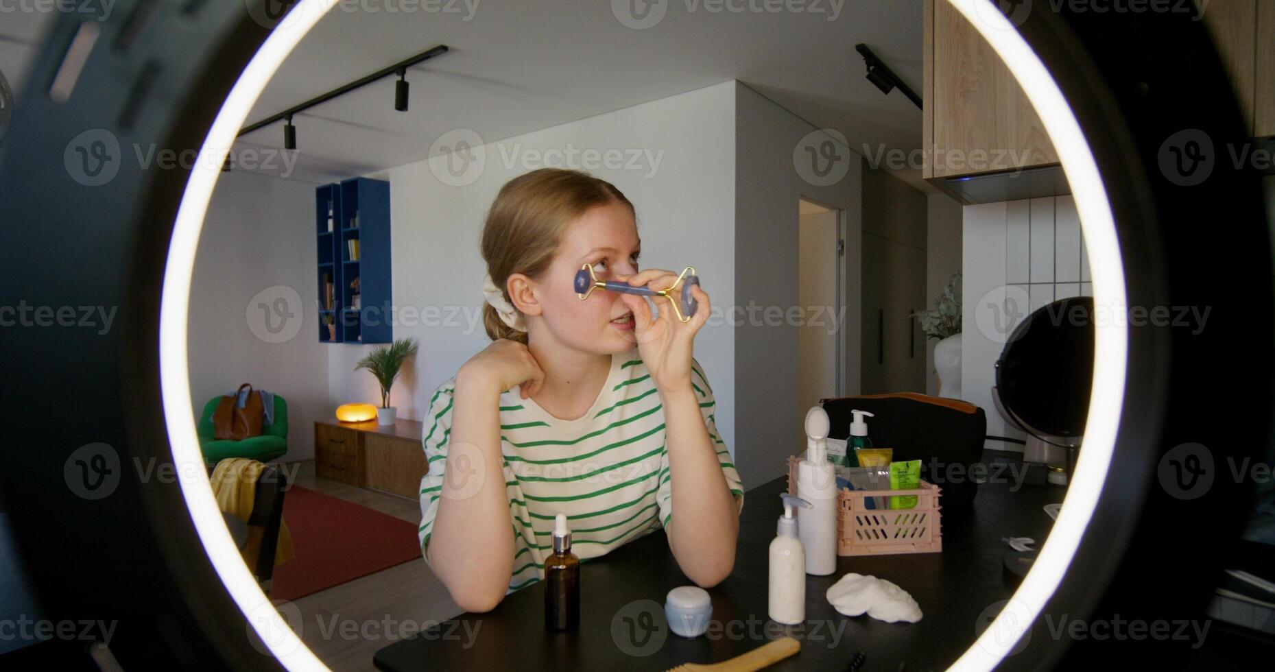 A young woman is doing a facial massage using a roller massager. She's talking while looking at the camera. through a round lamp photo