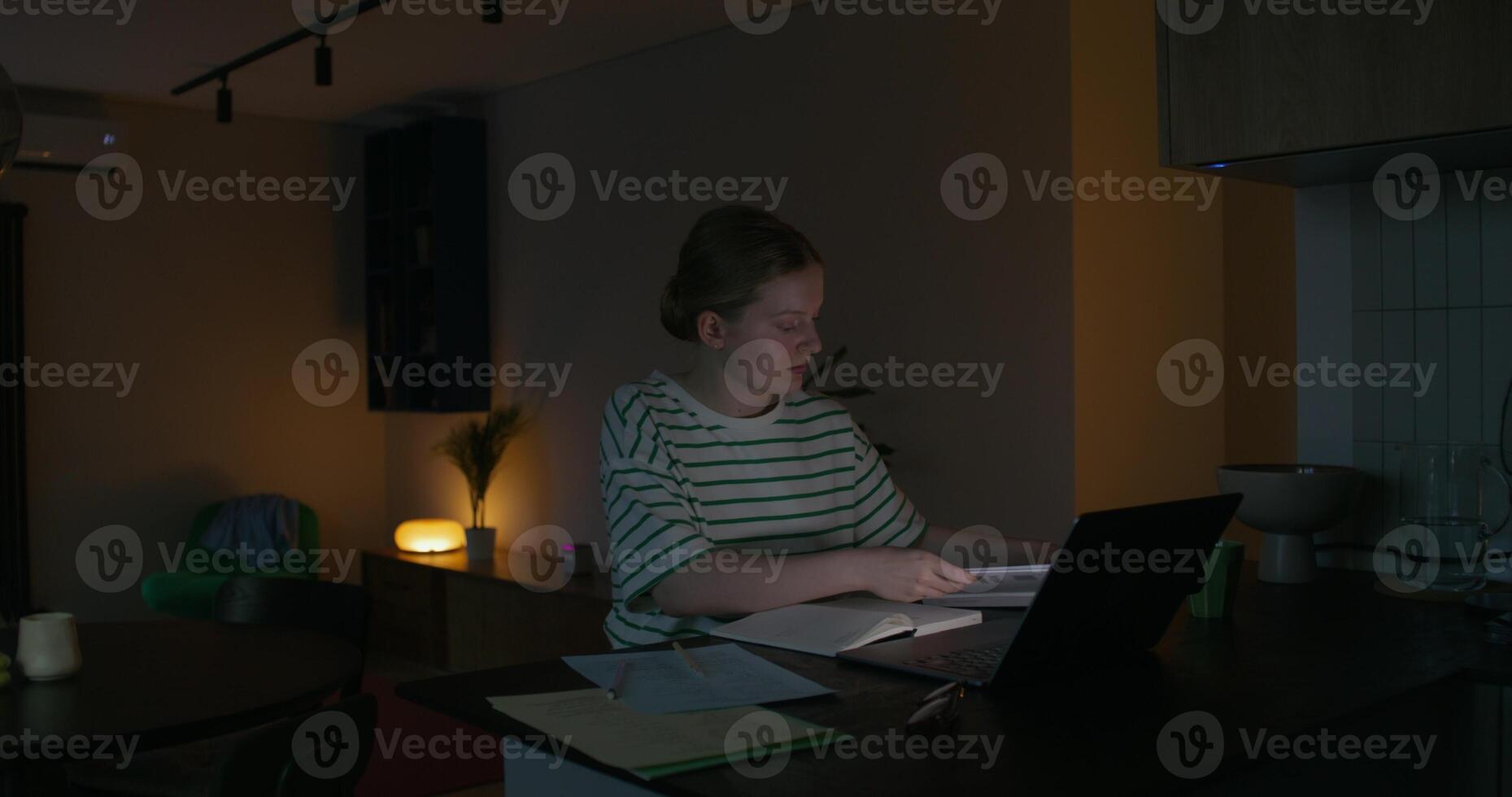 A woman is reading a book and writing in a notebook while sitting at a table in front of an open laptop. Dark room, home interior photo