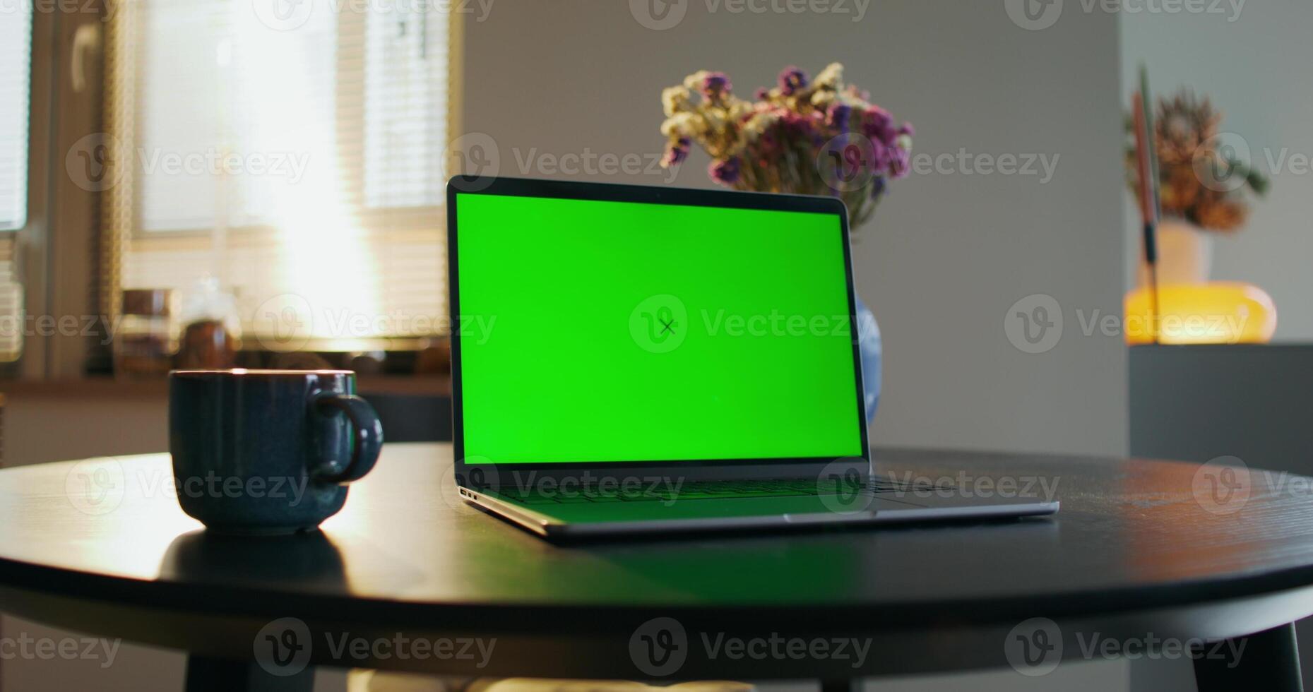 An open laptop with a green screen is on the table in the kitchen at home. A cup of hot drink nearby photo