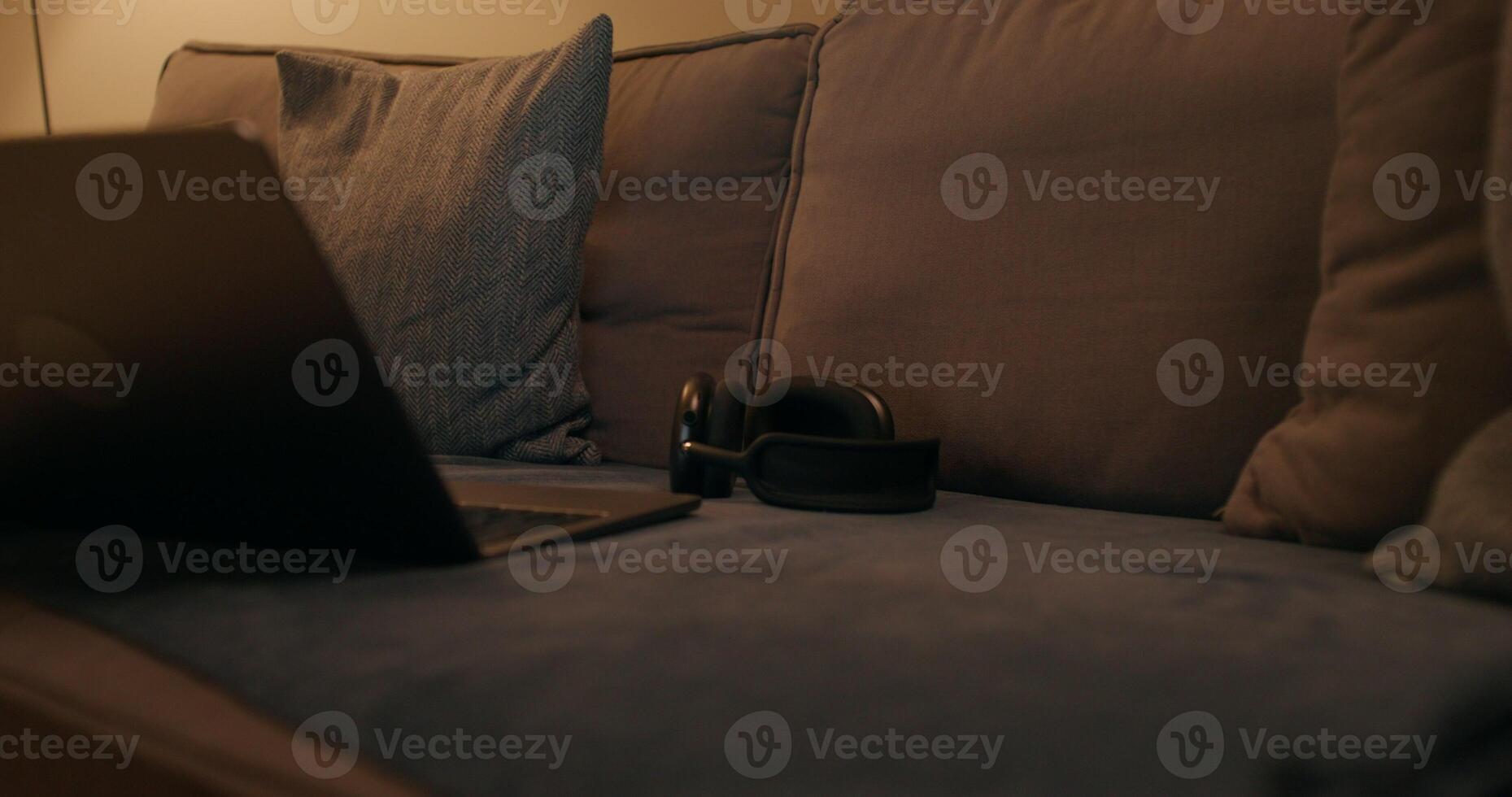 An open laptop and headphones are on the couch in the apartment, the evening light is dimmed, there are no people photo