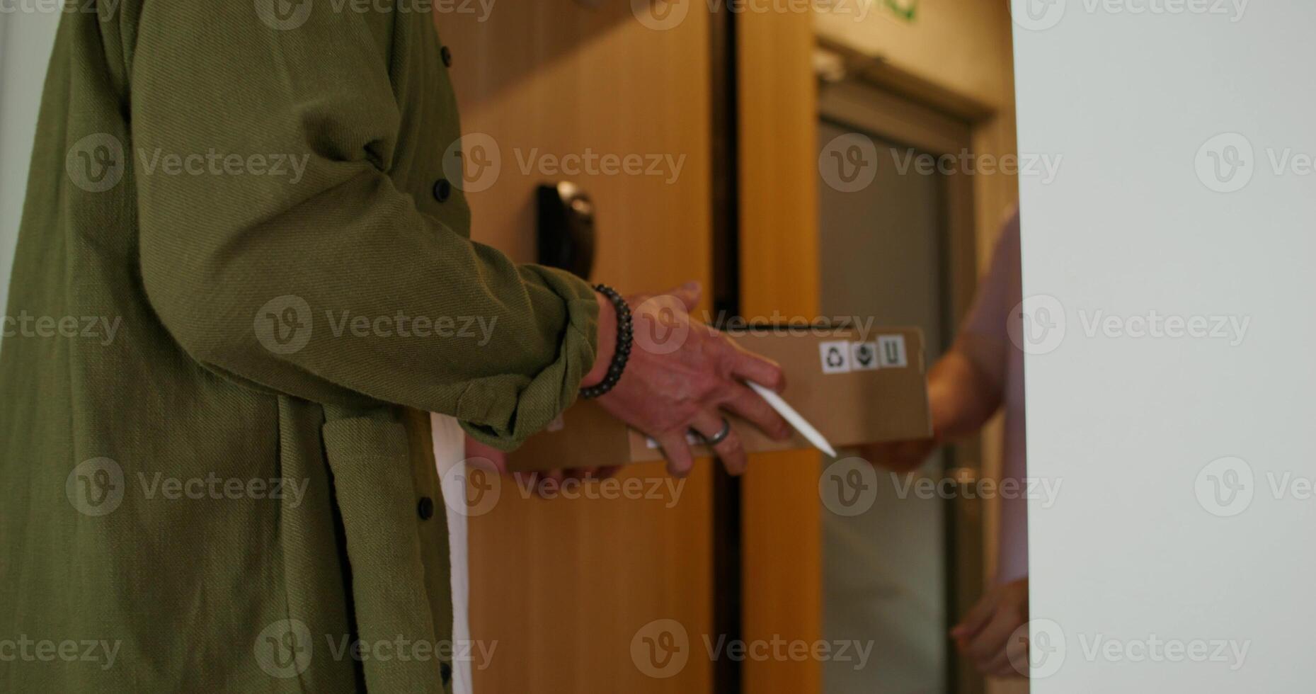 A man in casual clothes opens the front door and takes the box from the delivery guy. He signs with a stylus on the tablet. Daylight photo
