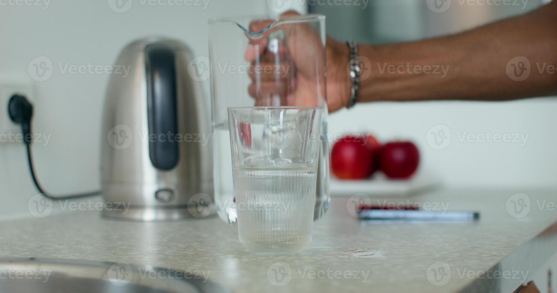 A man pours water from a carafe into a glass, close-up of his hands. Home interior, daylight photo