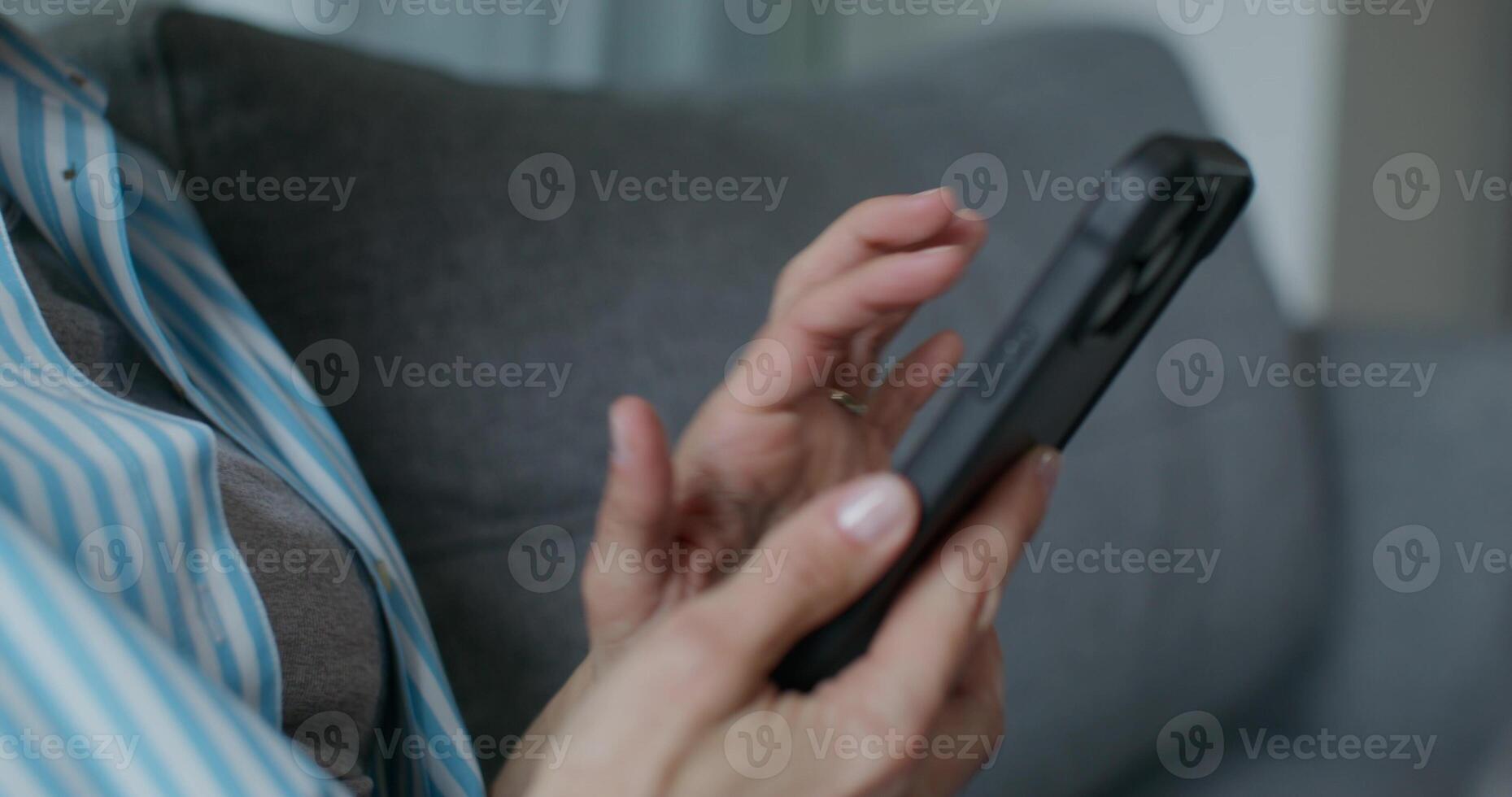 A woman in casual clothes uses a mobile phone while sitting on the couch at home. Close-up of her hands, an unrecognizable person photo