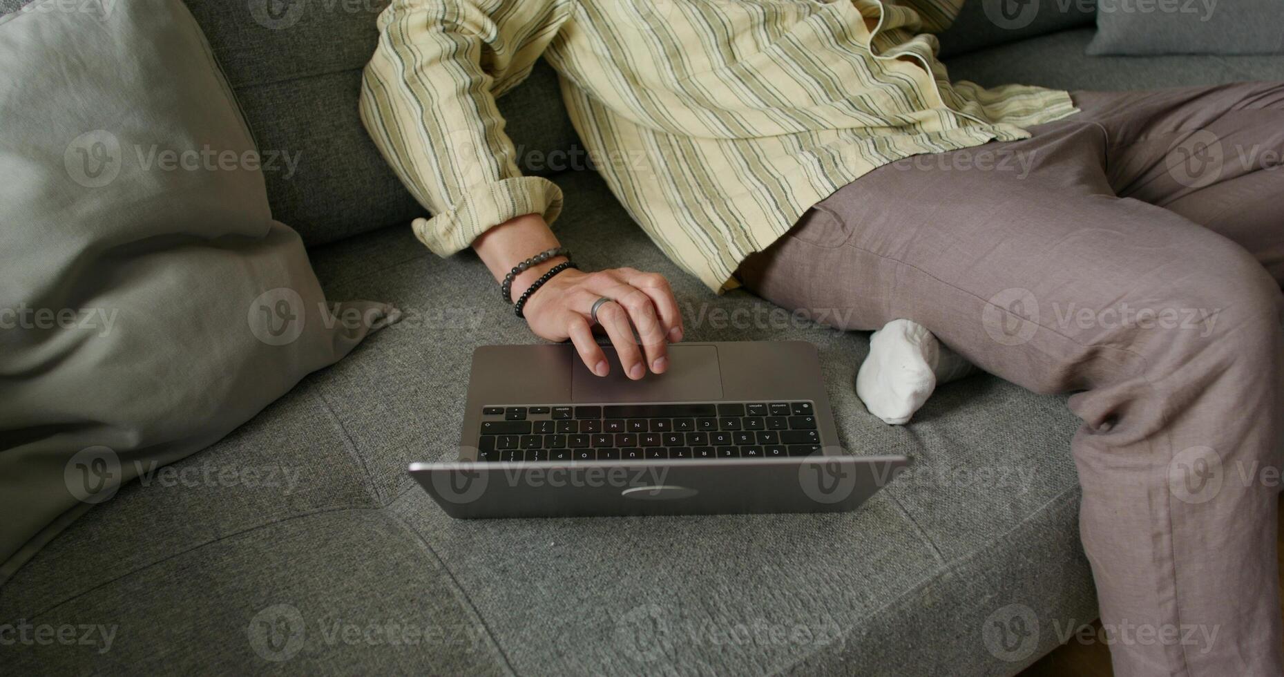 A man in casual clothes uses a laptop sitting on the couch at home. close-up of his hand, an unrecognizable person. Daylight photo