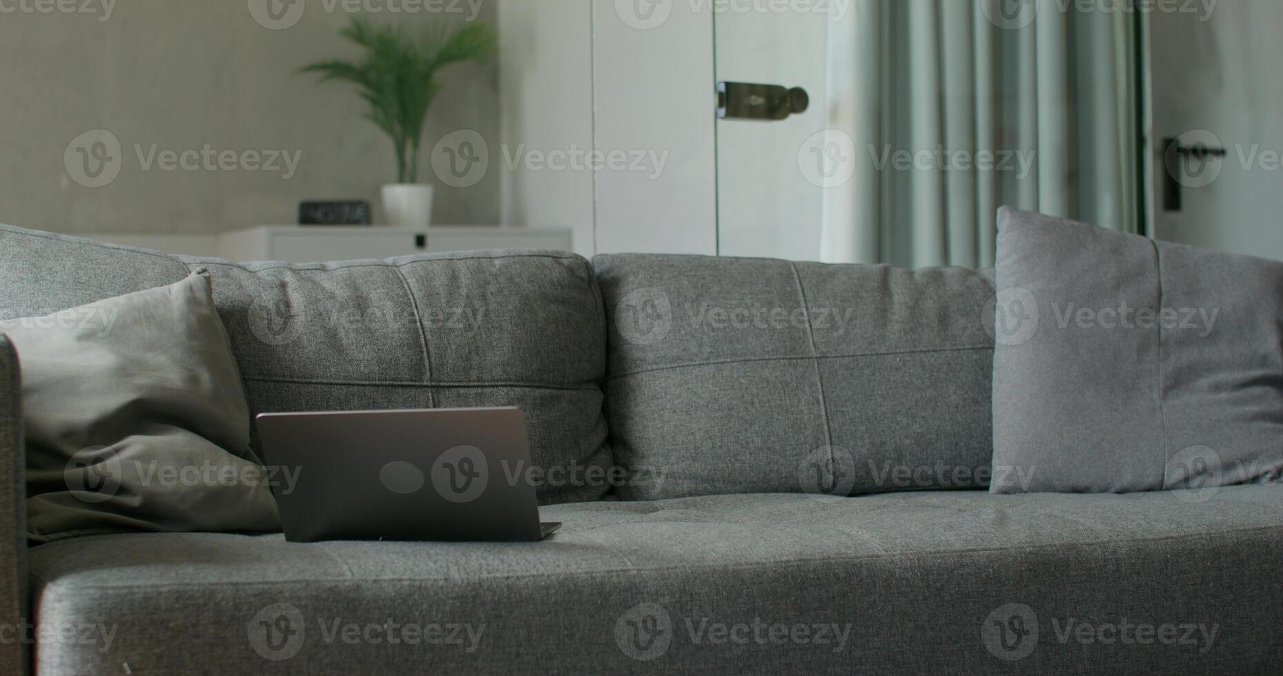 A bearded man in casual clothes sits down on the sofa and starts using his mobile phone and laptop. Daylight photo