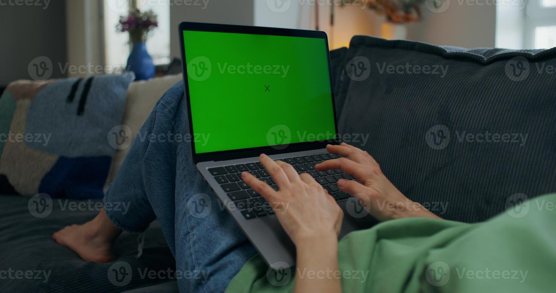 A woman typing on a laptop with green screen lying on the couch at home, close-up of her hands, an unrecognizable person photo