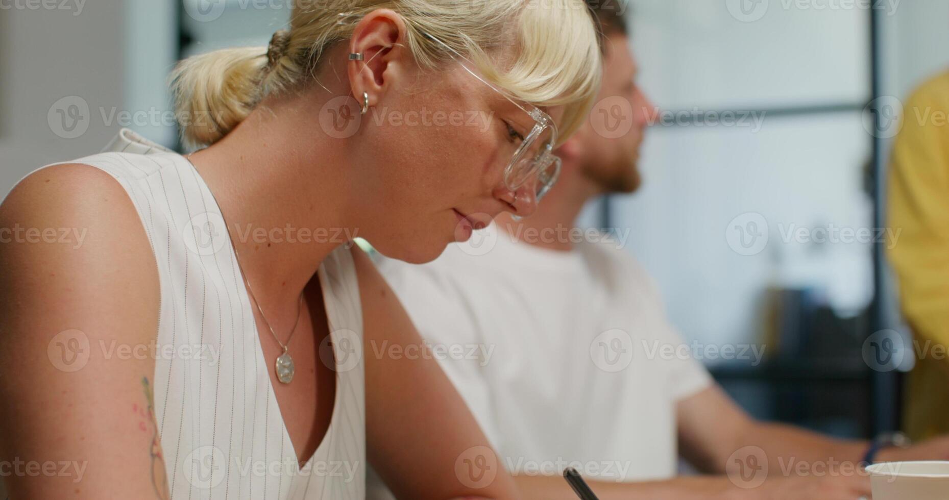 A woman makes notes in a document while sitting at an office meeting next to her colleagues. Daylight photo