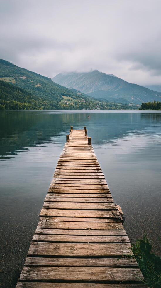Tranquil Wooden Dock Extends Over Calm Lake Surrounded by Mountains on a Cloudy Day photo