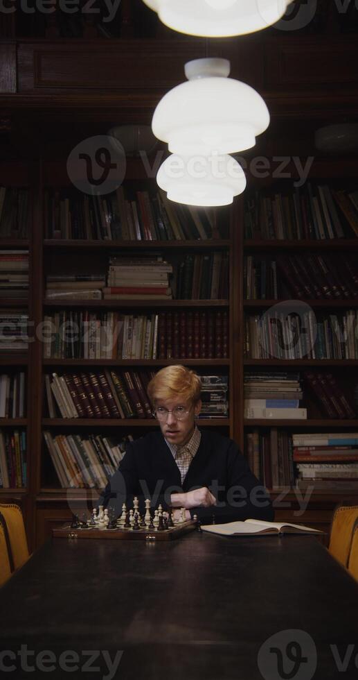 A young man of European appearance, dressed in a business suit, is studying with concentration to play chess alone, using a textbook, sitting at a table in the library, vertical shooting - business photo