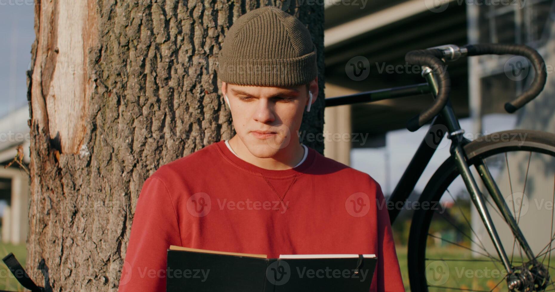 A young man of European appearance in a knitted hat reads a book and eats a sandwich, sitting on the ground under a tree next to the bridge. The bike stands under the tree next to the guy photo