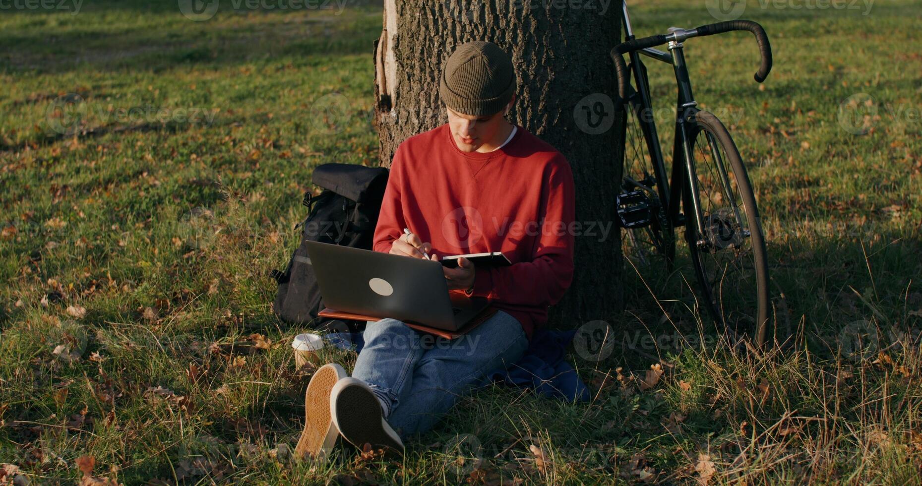 A young man of European appearance in a knitted hat uses a graphics tablet and a laptop, sitting on the ground under a tree. The bike stands next to the guy - graphics photo