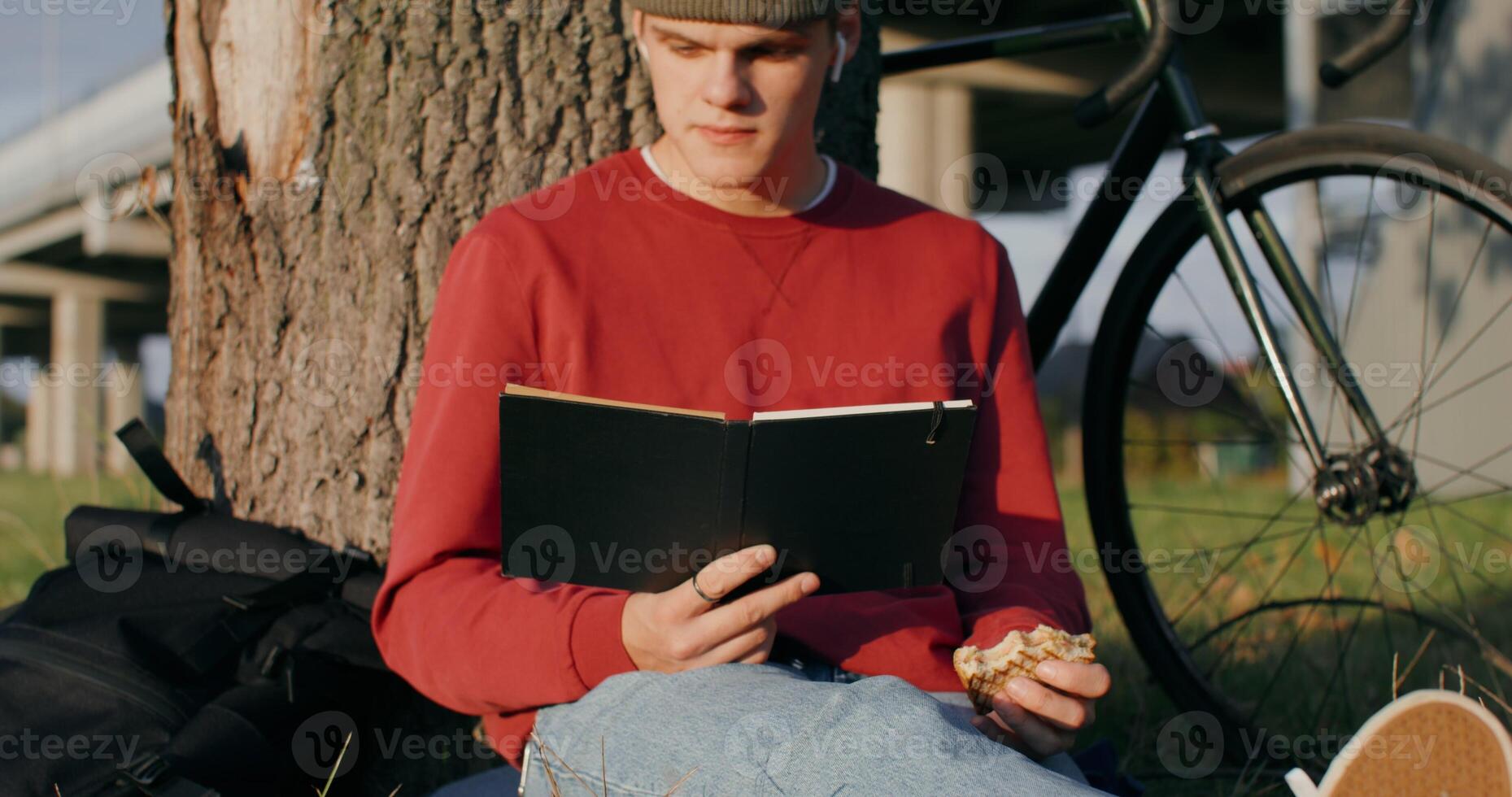 A young man of European appearance in a knitted hat reads a book and eats a sandwich, sitting on the ground under a tree next to the bridge. The bike stands under the tree next to the guy - knitted photo