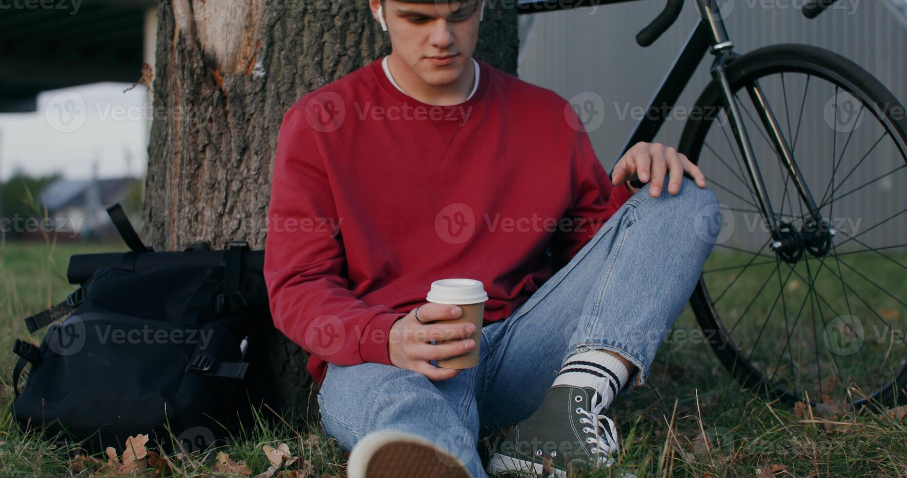 A young man of European appearance in a knitted hat and with wireless headphones drinks coffee while sitting on the ground under a tree near a bicycle, standing nearby - knitted photo