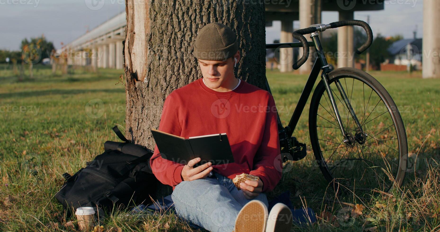 A young man of European appearance in a knitted hat reads a book and eats a sandwich, sitting on the ground under a tree next to the bridge. The bike stands under the tree next to the guy - sitting photo