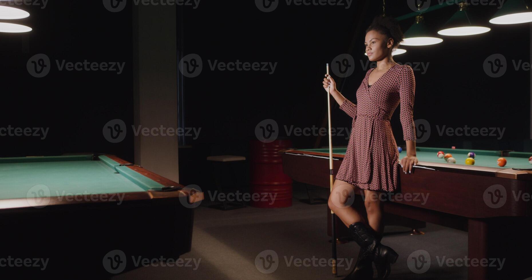 A beautiful young African-American woman, dressed in a dress, with a cue in her hands, stands leaning on a billiard table and smiles while looking at the camera photo