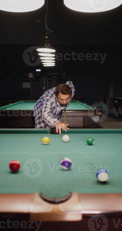 A young man of European appearance with a beard, dressed in casual clothes, plays billiards alone under dim lights in a billiard club, vertical photo