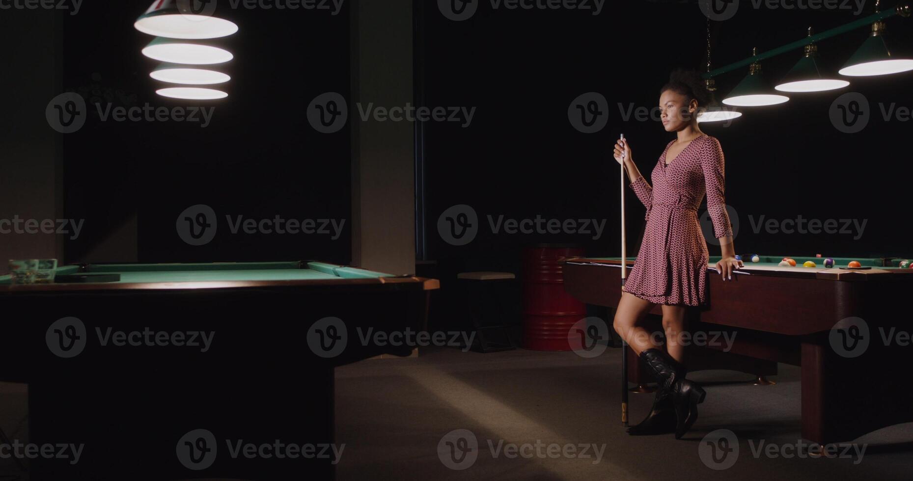 A beautiful young African-American woman, dressed in a dress, with a cue in her hands, stands leaning on a billiard table and smiles while looking at the camera - stands photo
