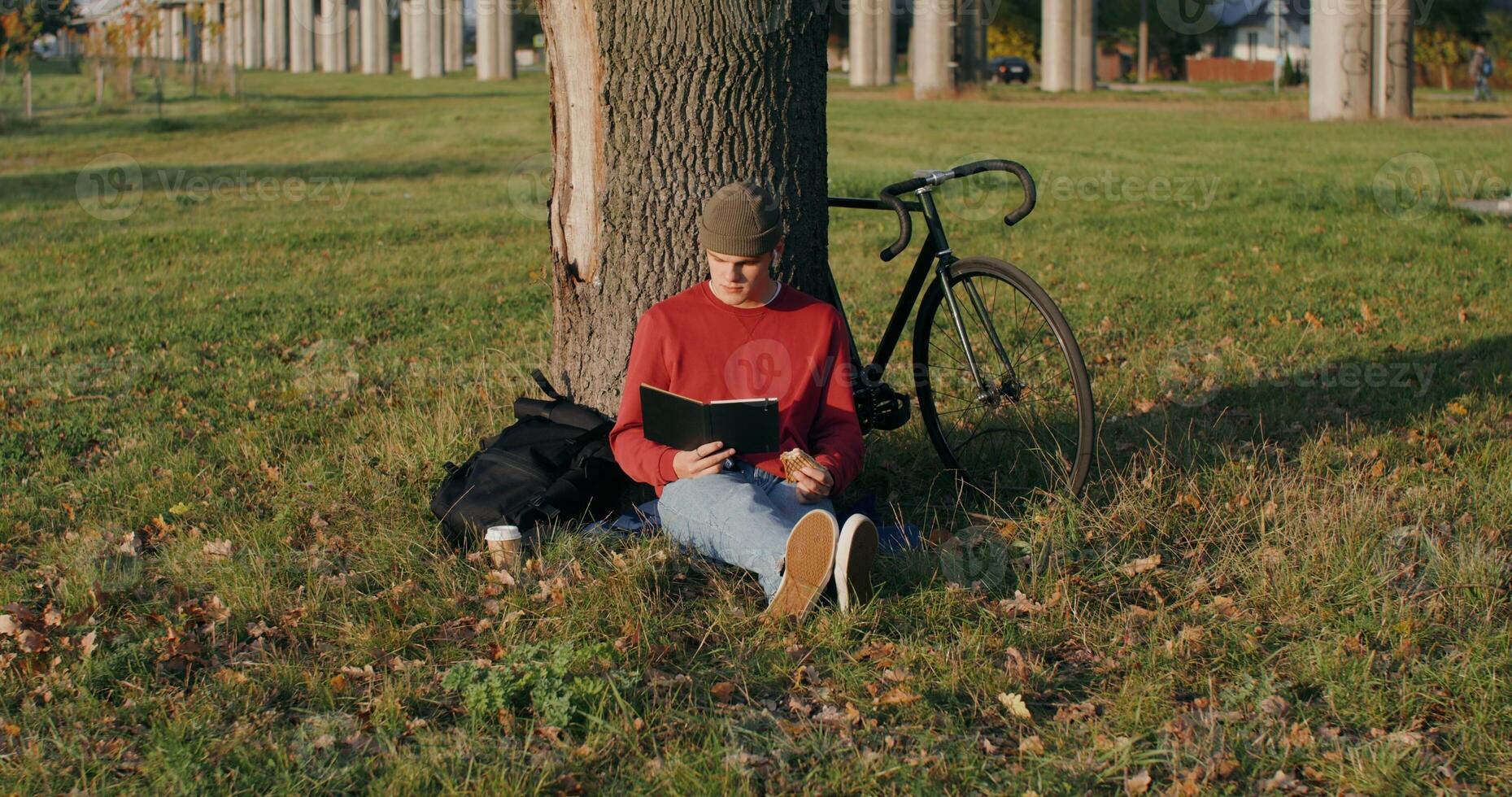 A young man of European appearance in a knitted hat reads a book and eats a sandwich, sitting on the ground under a tree next to the bridge. The bike stands under the tree next to the guy - ground photo