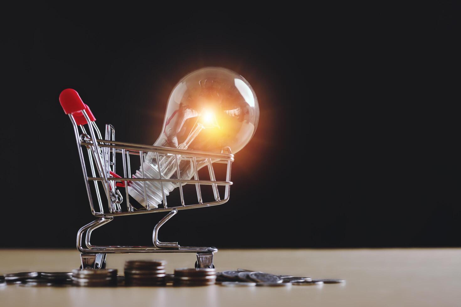 Light bulb idea is in the shopping cart and there are Money, coins and some stationery on wood table stack and on Black background in at home, Front view with copy space.New normal shopping concept. photo