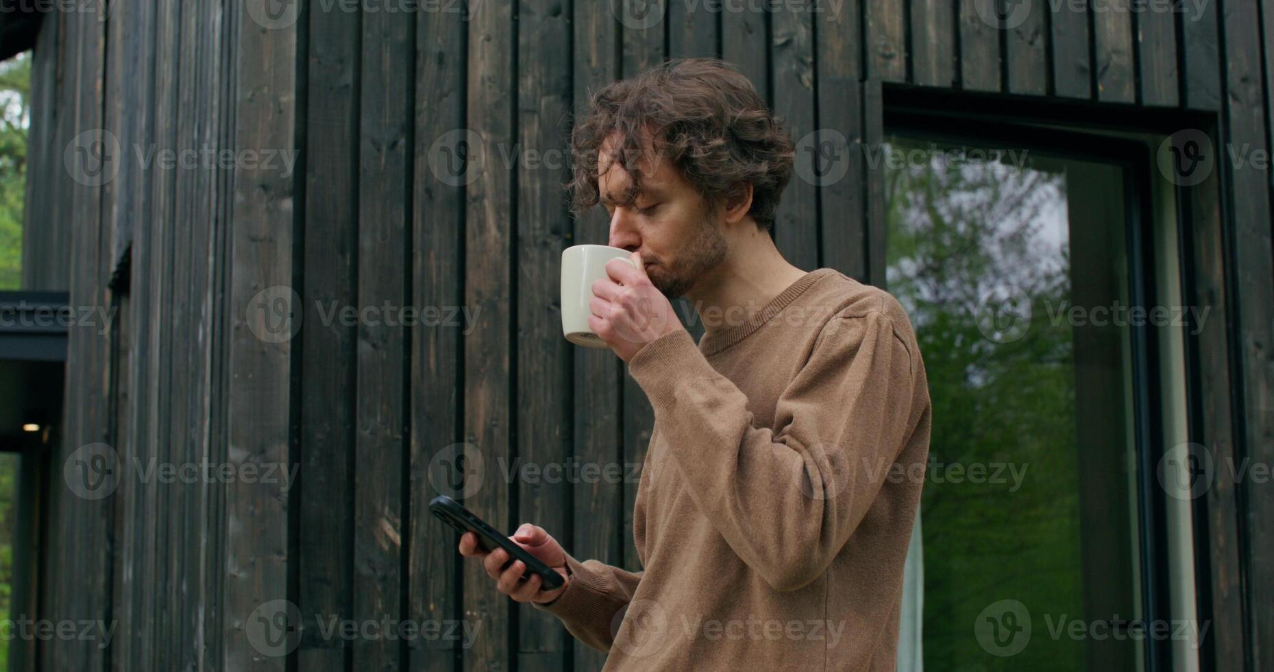 A man in casual clothes uses a mobile phone and drinks coffee standing on the terrace on a summer day. photo