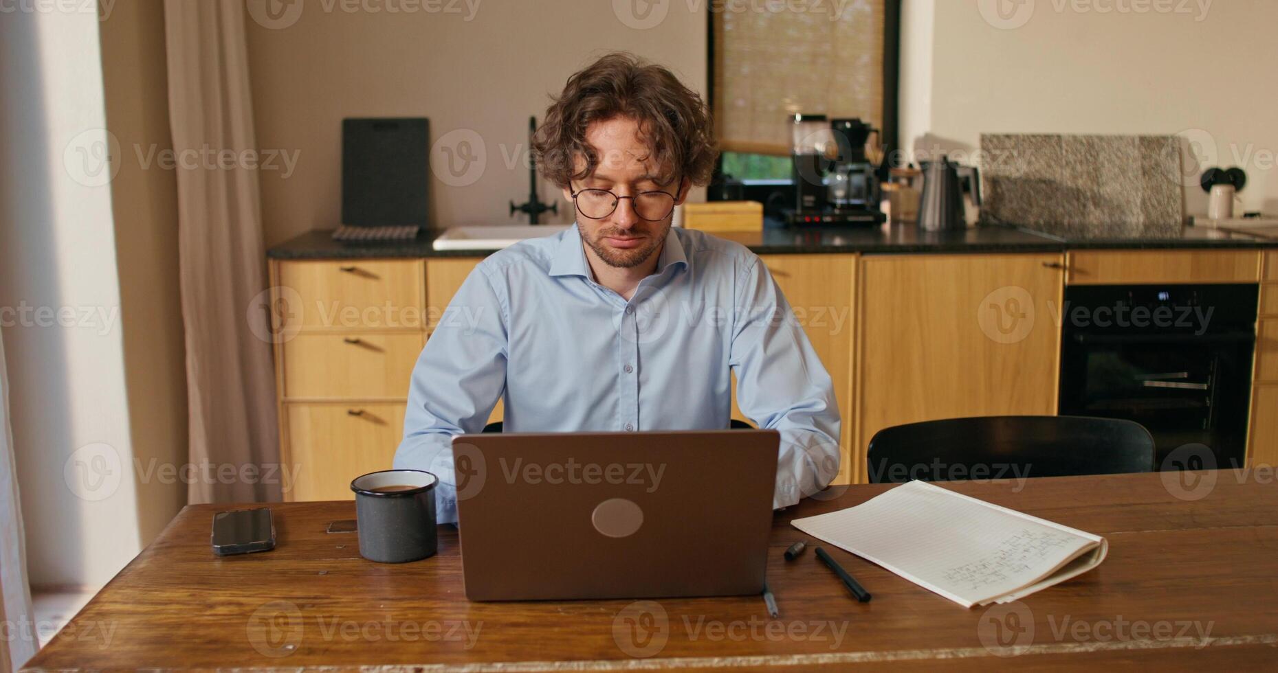 A man with glasses works from home. He uses a laptop and drinks coffee sitting at the kitchen table photo