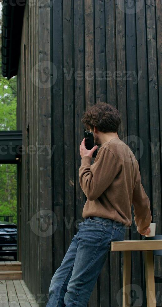 A man in casual clothes is talking on a mobile phone and drinking coffee standing on the terrace of a stylish cottage on a summer day. photo