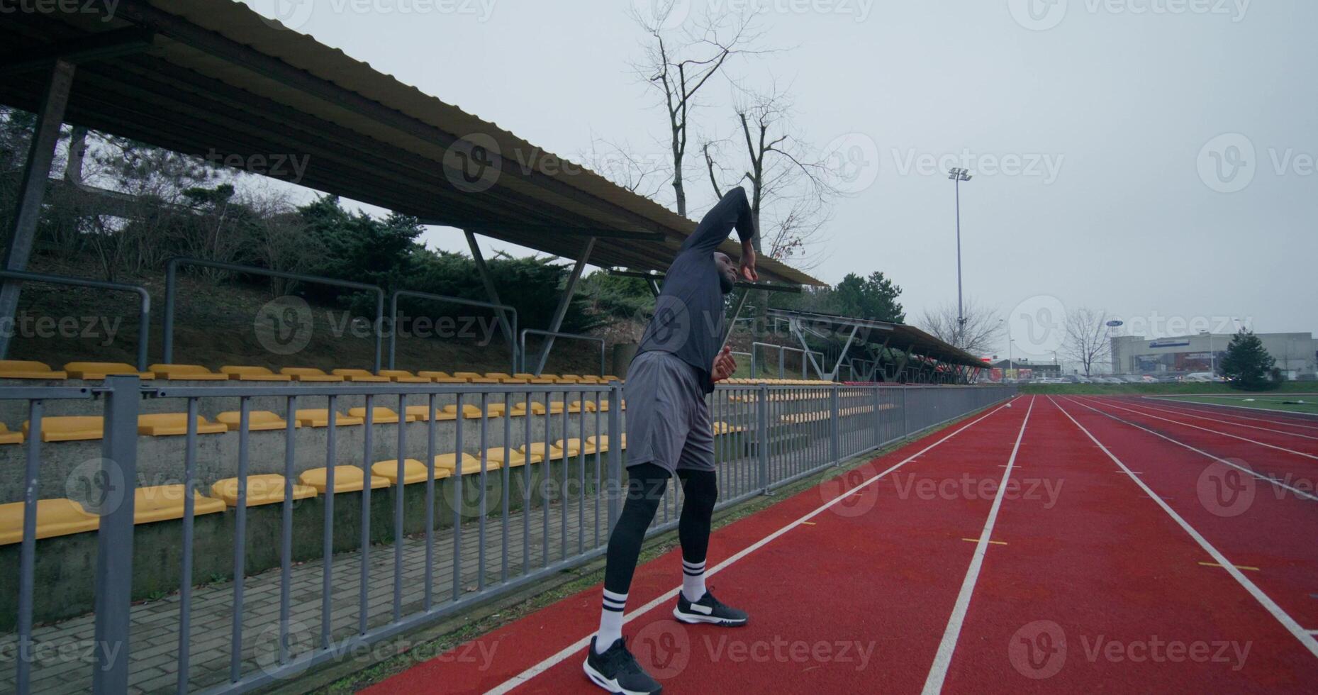 A man in a sports outfit with earphones in his ears does a warm-up standing at the stadium on a cold day - outfit photo