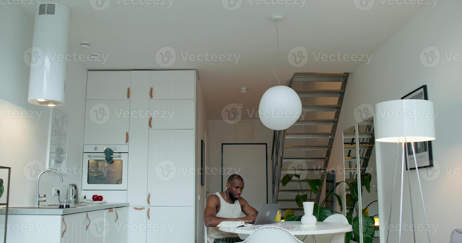 A man in pajamas uses a laptop and writes in a notebook sitting at a table in the kitchen at home with a serious look - sitting photo