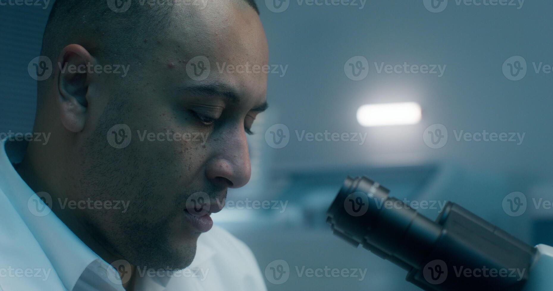 A man in a lab coat uses a microscope sitting at a desk in a medical laboratory. Close-up of his focused face - medical photo
