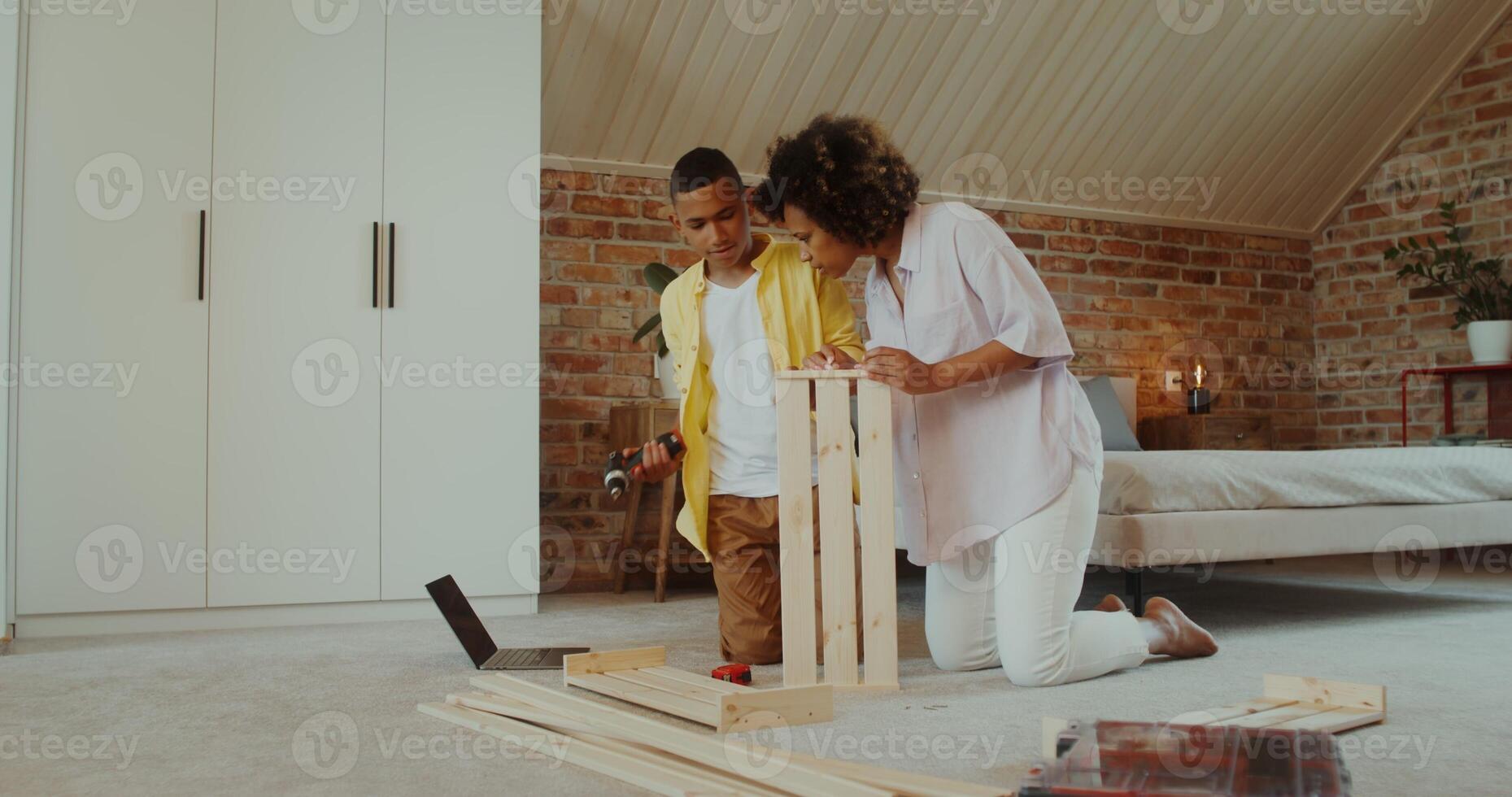 Mom and son make wooden furniture using a drill, sitting on the floor in front of an open laptop at home photo
