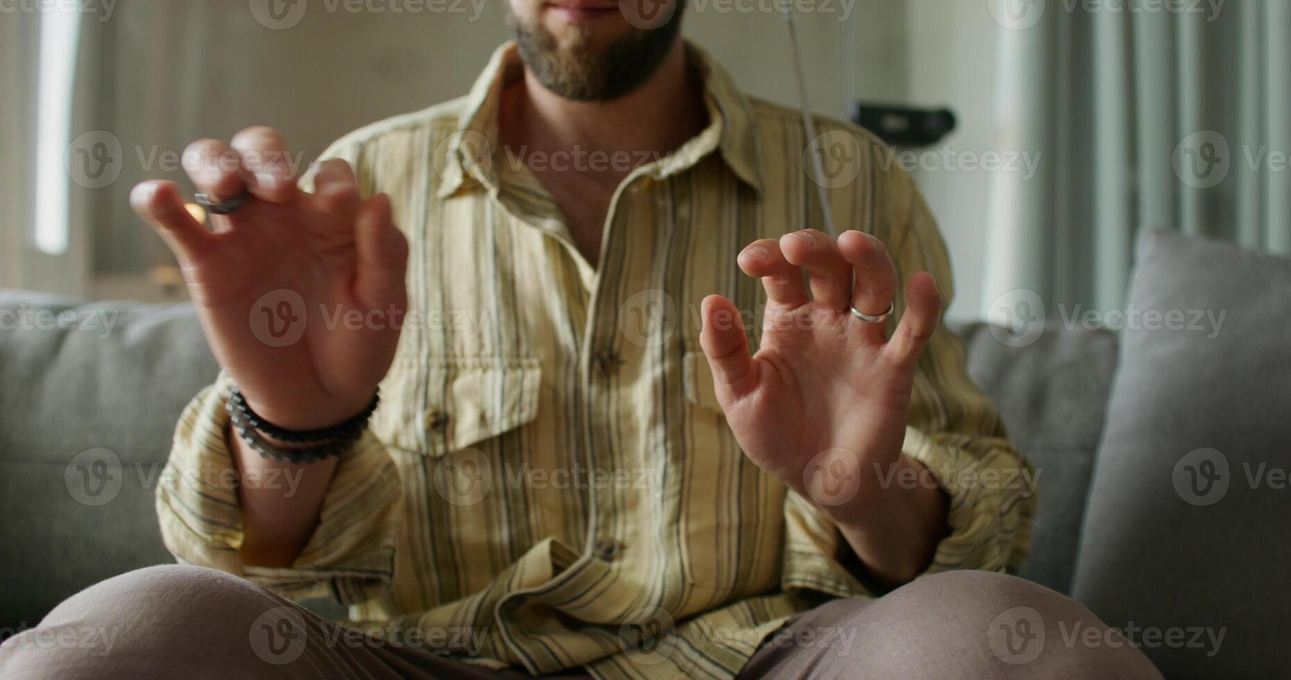 A man in a virtual reality headset is typing on a virtual keyboard and moving his hands sitting on the couch. Daylight, home interior photo