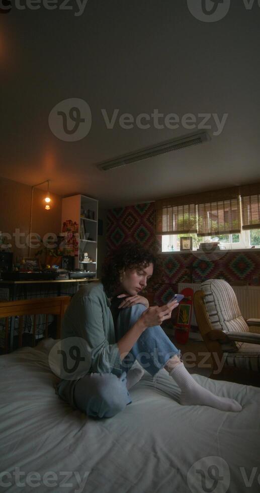 A young woman uses a mobile phone while sitting on a bed at home with a depressed look photo